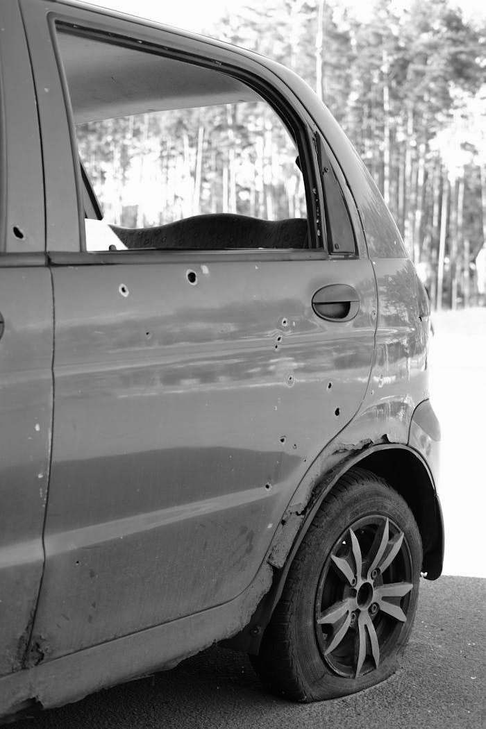 Close-up of a car door with bullet holes and a flat tire in a forest setting.